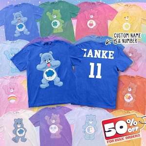 May include: A collection of colorful t-shirts featuring various Care Bear designs. The shirts are in shades of blue, green, pink, yellow, and orange. One shirt has the name "Zanke" and the number "11" on the back. The text "CUSTOM NAME & NUMBER" is also visible.