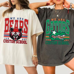 May include: Two oversized t-shirts. One is cream-colored with a red, white, and blue graphic of a bear wearing star-shaped sunglasses and the words "WE ARE BEARS CUSTOM SCHOOL." The other is dark gray with a green alligator graphic and the words "WE ARE GATORS CUSTOM SCHOOL."