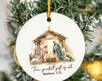 May include: White ceramic Christmas ornament with a Nativity scene illustration. The image depicts Mary, Joseph, and baby Jesus in a manger, with animals and the text "The greatest gift of all" and "Matthew 1:21". A gold cord is attached for hanging.