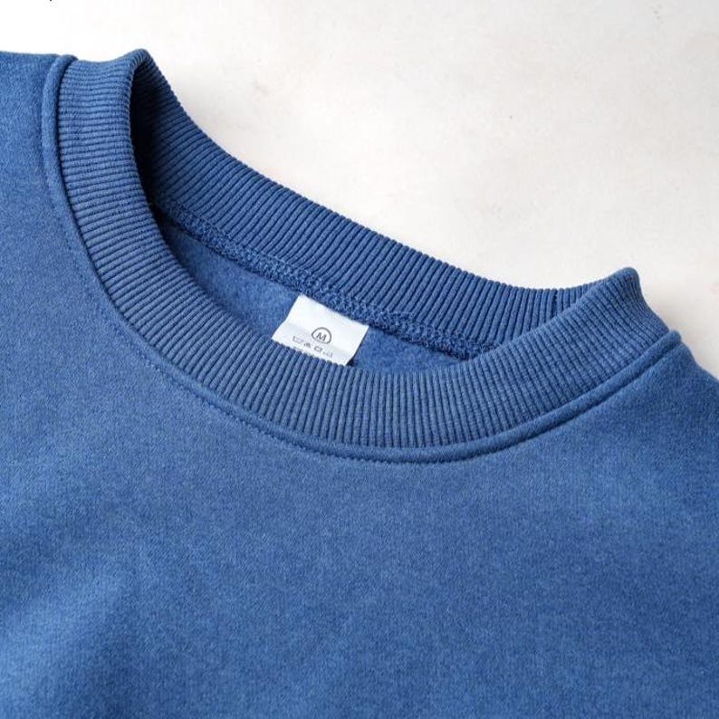 Peut inclure: Gros plan sur un sweat-shirt bleu &agrave; col rond. Le col et les poignets c&ocirc;tel&eacute;s sont d'un bleu plus fonc&eacute;. Une &eacute;tiquette blanche avec un petit logo est visible pr&egrave;s du col.