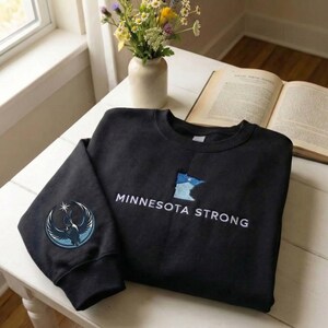 Peut inclure: Un sweat-shirt noir avec les mots "MINNESOTA STRONG" brod&eacute;s en blanc. Un contour bleu et blanc de l'&Eacute;tat du Minnesota est sur le devant. Un &eacute;cusson circulaire avec un motif d'oiseau est sur la manche gauche.