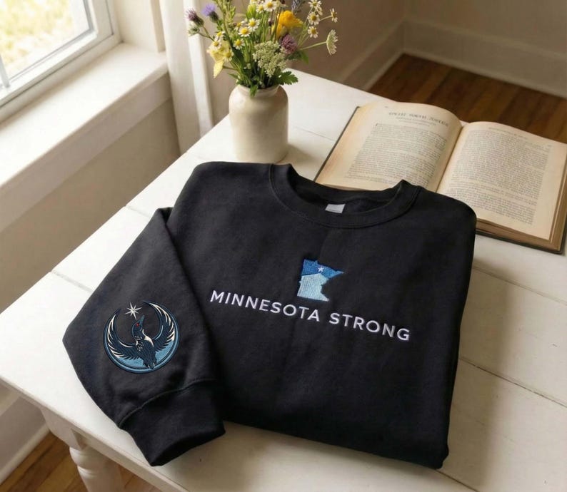 Peut inclure: Un sweat-shirt noir avec les mots "MINNESOTA STRONG" brod&eacute;s en blanc. Un contour bleu et blanc de l'&Eacute;tat du Minnesota est sur le devant. Un &eacute;cusson circulaire avec un motif d'oiseau est sur la manche gauche.