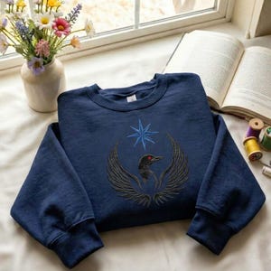 Puede incluir: Sudadera azul marino con un dise&ntilde;o bordado de un p&aacute;jaro con las alas extendidas, un ojo rojo y una estrella azul. La sudadera se muestra sobre una superficie blanca cerca de una ventana, con un jarr&oacute;n de flores y un libro abierto en el fondo.