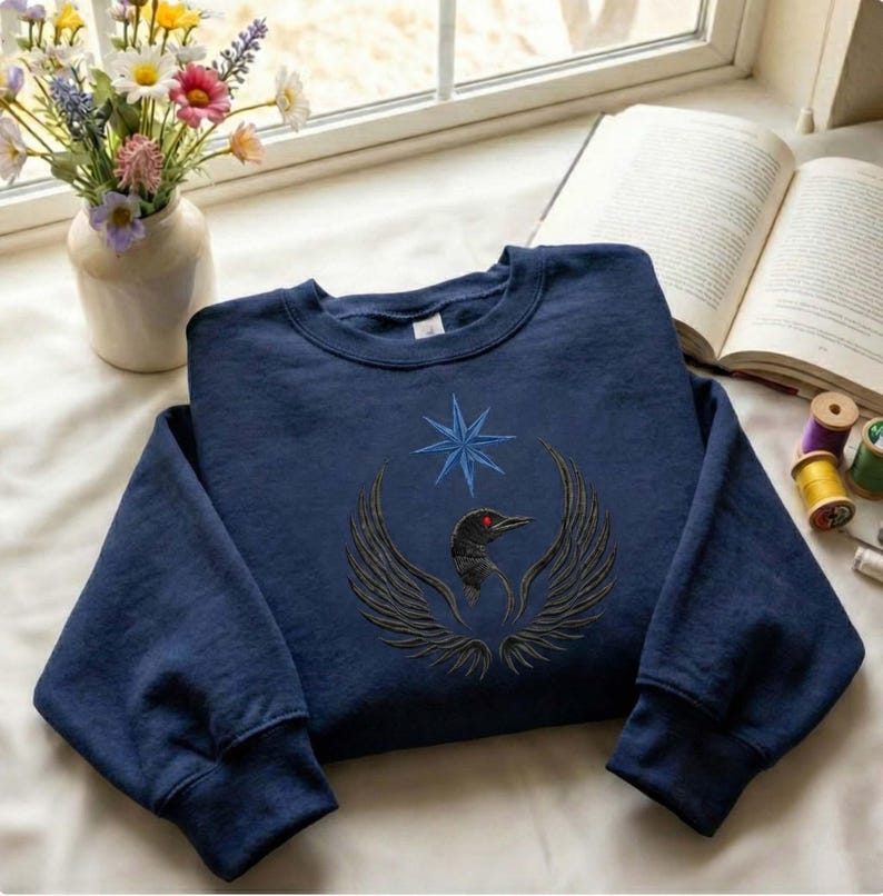 Puede incluir: Sudadera azul marino con un dise&ntilde;o bordado de un p&aacute;jaro con las alas extendidas, un ojo rojo y una estrella azul. La sudadera se muestra sobre una superficie blanca cerca de una ventana, con un jarr&oacute;n de flores y un libro abierto en el fondo.