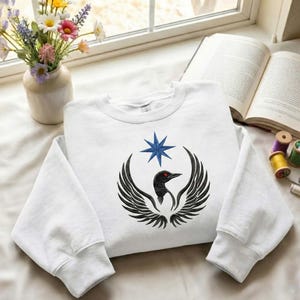 Puede incluir: Sudadera blanca con un dise&ntilde;o de p&aacute;jaro negro con alas extendidas, un ojo rojo y una estrella azul. El dise&ntilde;o est&aacute; centrado en la parte delantera de la sudadera, doblada sobre una superficie blanca.