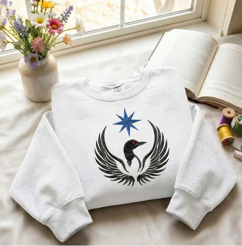 Puede incluir: Sudadera blanca con un dise&ntilde;o de p&aacute;jaro negro con alas extendidas, un ojo rojo y una estrella azul. El dise&ntilde;o est&aacute; centrado en la parte delantera de la sudadera, doblada sobre una superficie blanca.