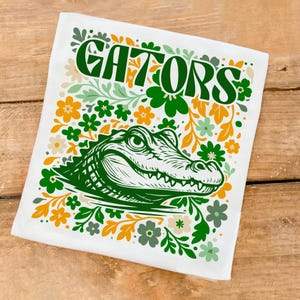 May include: White rectangular decorative towel with the word "GATORS" in green, surrounded by a green alligator illustration and floral design in green, orange, and cream colors.