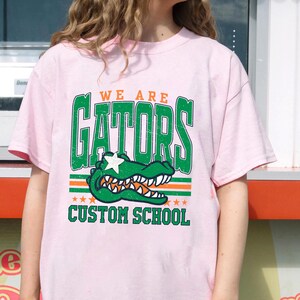 May include: A light pink t-shirt featuring a green alligator graphic with the text "WE ARE GATORS" above and "CUSTOM SCHOOL" below. The alligator has a white star on its head and is set against orange and green stripes.