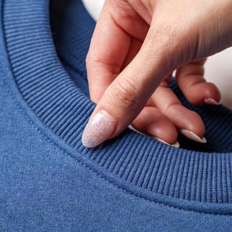 Peut inclure: Gros plan sur un sweat-shirt bleu avec un col c&ocirc;tel&eacute;. Le tissu semble doux et la couleur est une nuance de bleu profond. Une main avec du vernis &agrave; ongles paillet&eacute; touche le col.