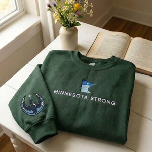 Peut inclure: Un sweat-shirt vert fonc&eacute; avec le texte "MINNESOTA STRONG" en blanc. Le sweat-shirt pr&eacute;sente un &eacute;cusson de l'&Eacute;tat du Minnesota en bleu et blanc, et un &eacute;cusson circulaire avec un motif d'oiseau sur la manche.