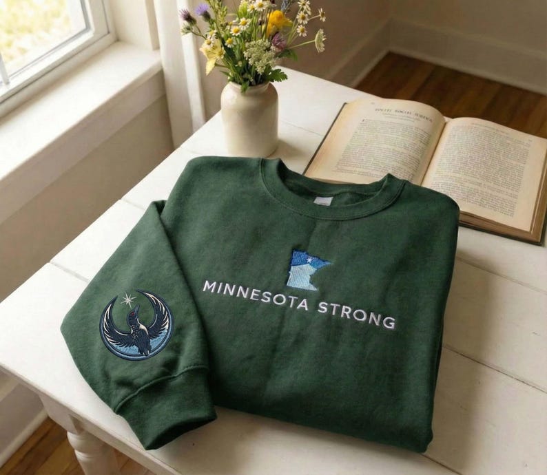 Peut inclure: Un sweat-shirt vert fonc&eacute; avec le texte "MINNESOTA STRONG" en blanc. Le sweat-shirt pr&eacute;sente un &eacute;cusson de l'&Eacute;tat du Minnesota en bleu et blanc, et un &eacute;cusson circulaire avec un motif d'oiseau sur la manche.