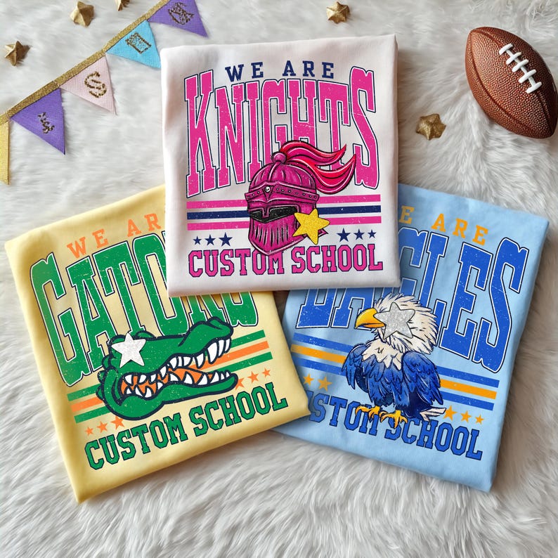 May include: Three custom school t-shirts in yellow, white, and blue. The white shirt says "We Are Knights" with a knight's helmet graphic. The yellow shirt says "We Are Gators" with an alligator graphic. The blue shirt says "We Are Eagles" with an eagle graphic.