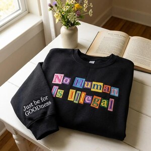 Peut inclure: Sweat-shirt noir avec l'inscription "No Human Is Illegal" en lettres color&eacute;es. La manche porte l'inscription "Just be for GOODness" en blanc. Le sweat-shirt est pos&eacute; sur une table blanche, avec un vase de fleurs et un livre ouvert.