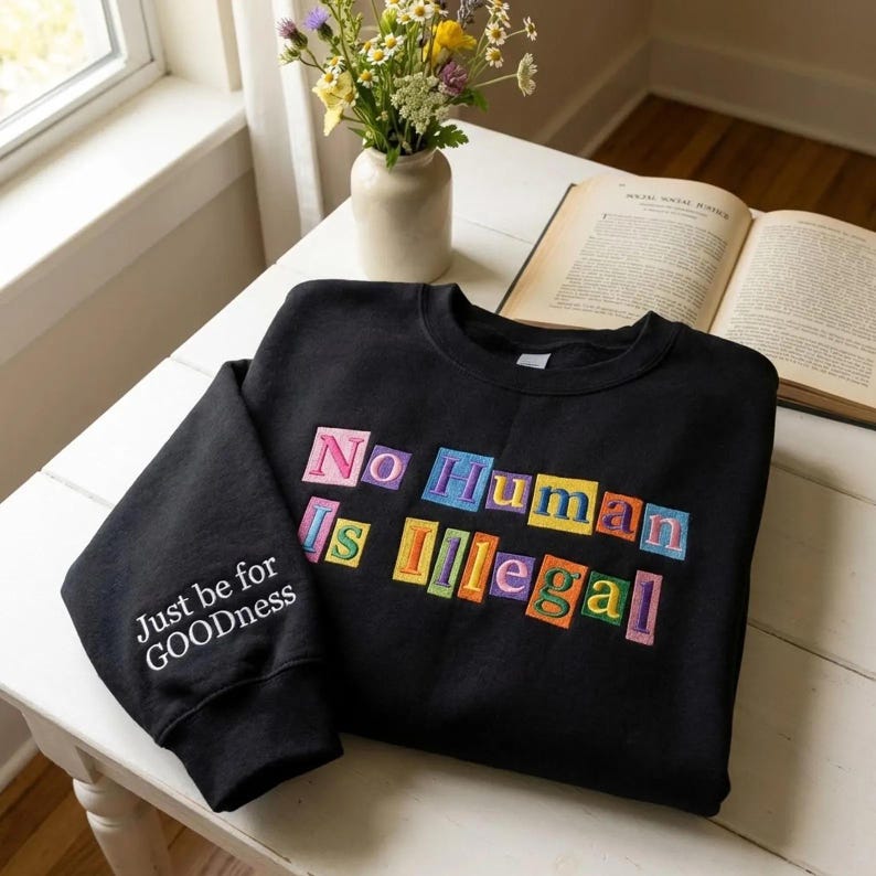 Peut inclure: Sweat-shirt noir avec l'inscription "No Human Is Illegal" en lettres color&eacute;es. La manche porte l'inscription "Just be for GOODness" en blanc. Le sweat-shirt est pos&eacute; sur une table blanche, avec un vase de fleurs et un livre ouvert.