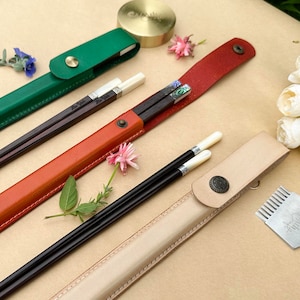 K&ouml;nnte beinhalten: Set aus drei Leder-Essst&auml;bchen-Etuis in Gr&uuml;n, Orange und Beige. Die Etuis sind geschlossen und haben einen dekorativen Knopf. Die Essst&auml;bchen sind im orangen Etui sichtbar. Die Essst&auml;bchen sind schwarz mit wei&szlig;en Spitzen und haben ein dekoratives Design.