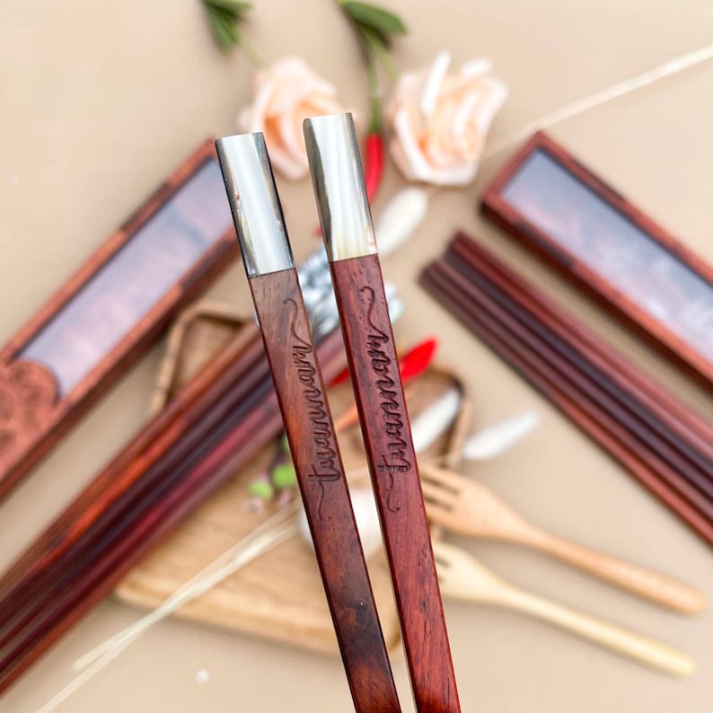Puede incluir: Un par de palillos de madera rojos con puntas plateadas. Los palillos est&aacute;n grabados con las palabras "In Love We Trust" y "Forever and Always".