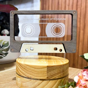 Puede incluir: Una luz nocturna con forma de casete de madera con un inserto de acrílico transparente. El casete tiene una etiqueta blanca con el texto "Scan QR code music of Signature". La luz nocturna está sobre una base de madera con un botón de encendido.
