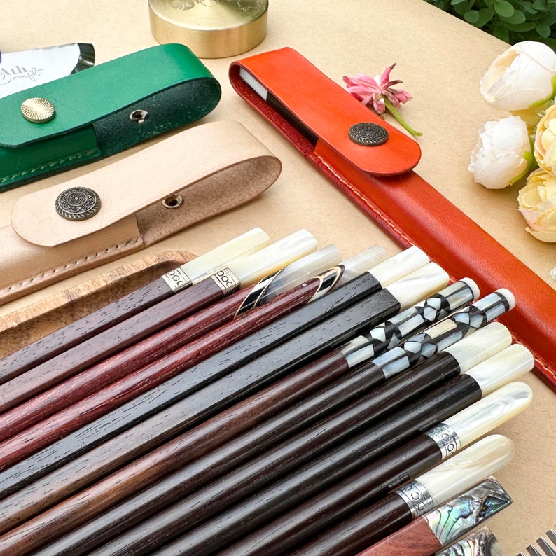 K&ouml;nnte beinhalten: Ein Satz Holzst&auml;bchen mit Perlmutteinlagen. Die St&auml;bchen befinden sich in einem braunen Lederetui mit einem Metallverschluss. Die St&auml;bchen sind f&auml;cherf&ouml;rmig angeordnet, wobei die Spitzen der St&auml;bchen nach oben zeigen.