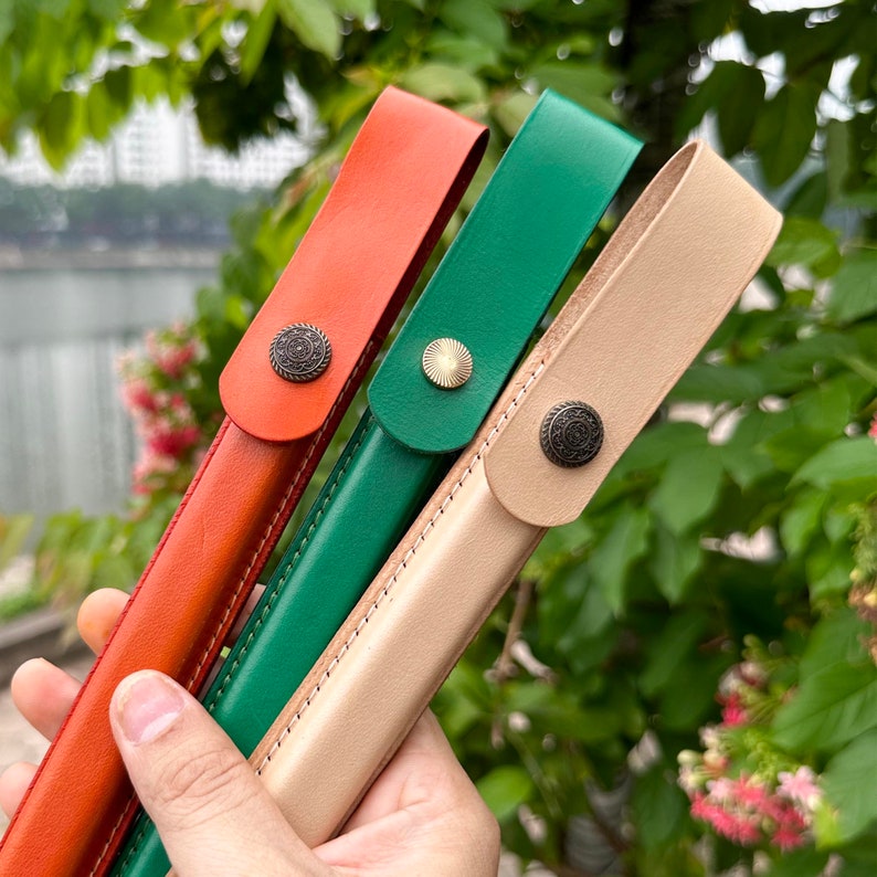 K&ouml;nnte beinhalten: Drei Leder-Stift-Etuis in Orange, Gr&uuml;n und Beige. Jedes Etui hat einen Metallknopfverschluss mit einem dekorativen Design.