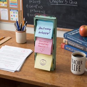 May include: A vertical wooden note holder with three stacked sticky notes. The top note is light blue with "Don't worry be happy!" written in purple. The middle note is pink with "You are a AMAZING Teacher !!!" and a heart. The bottom note is yellow with a smiley face.