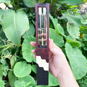 Puede incluir: Una caja de madera con una ventana de pl&aacute;stico transparente, que muestra tres palillos de madera. La caja est&aacute; decorada con un dise&ntilde;o floral en rojo y negro.