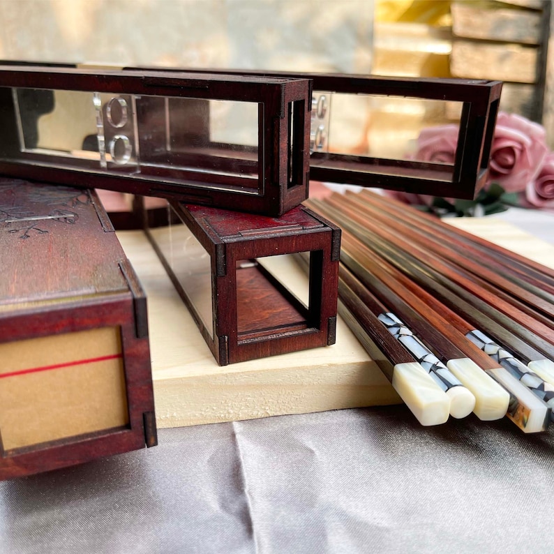 Puede incluir: Un juego de palillos de madera con puntas blancas decorativas, presentados en una caja de madera con tapa de acr&iacute;lico transparente. La caja es de madera oscura con veta de madera natural.