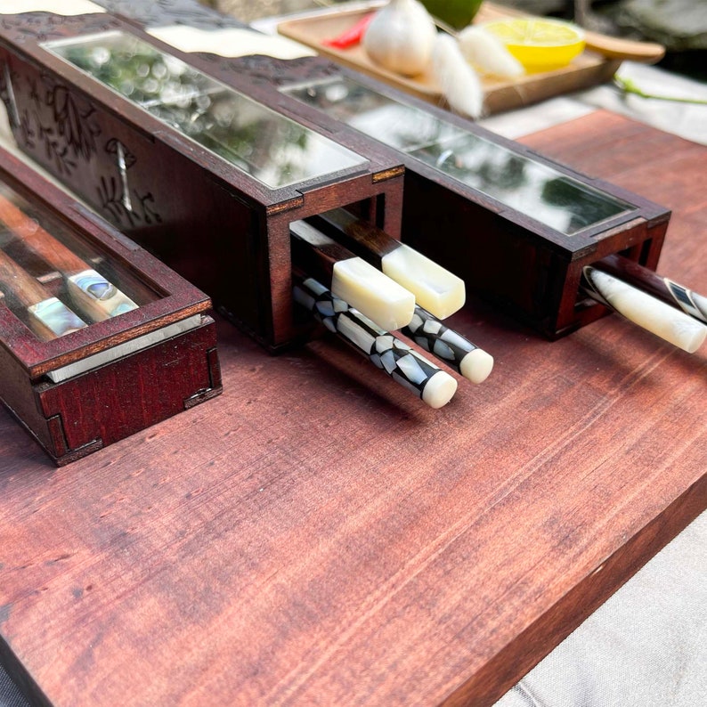 Puede incluir: Dos cajas de madera con tapas de vidrio, cada una contiene un par de palillos. Los palillos son de madera oscura con detalles blancos y negros. Las cajas est&aacute;n decoradas con tallas intrincadas.