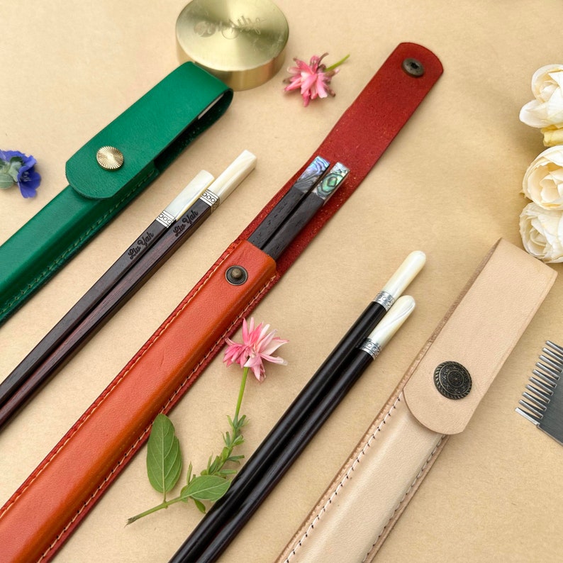 K&ouml;nnte beinhalten: Vier verschiedene Sets von Essst&auml;bchen in ihren Etuis. Die Etuis sind aus Leder und in Gr&uuml;n, Orange, Rot und Beige erh&auml;ltlich. Die Essst&auml;bchen sind aus Holz und haben dekorative Einlagen. Die Essst&auml;bchen sind mit "Bao Vah" beschriftet.