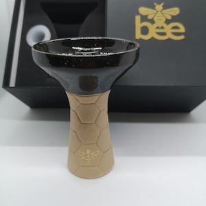Könnte beinhalten: Eine schwarze und beige Keramik-Shisha-Schale mit einem Wabenmuster und einem goldenen Bienenlogo. Die Schale befindet sich in einer schwarzen Box mit einem goldenen Bienenlogo.
