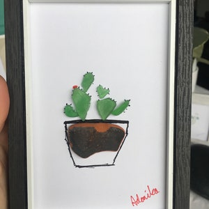 May include: A framed artwork featuring a cactus made of green and brown sea glass in a white pot with black outlines. The artwork is signed "Adonila" in red ink.