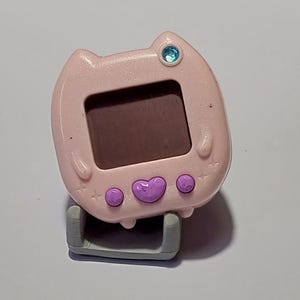 Puede incluir: Un juguete digital de mascota de mano en color rosa claro con forma de cabeza de gato, que incluye una pantalla cuadrada, tres botones morados y una pequeña gema azul. El dispositivo tiene orejas de gato moldeadas y descansa sobre un soporte gris.