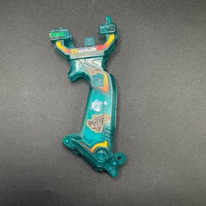 May include: A translucent teal plastic toy launcher with yellow and red detailing. The launcher features a handle and a top section with the word "BEYBLADE" printed on it. The toy is designed for launching spinning tops.