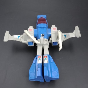 May include: A blue, gray, and white toy robot in a flying configuration. The toy has wings and a cockpit. The robot's legs are blue, and the arms and wings are gray. The toy is a vintage collectible.