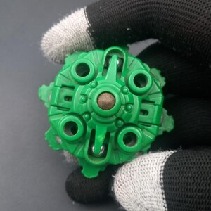 May include: A green plastic toy component with a circular design, featuring multiple holes and a central brown element. The object is held by a hand wearing a black and white glove, with a dark background.