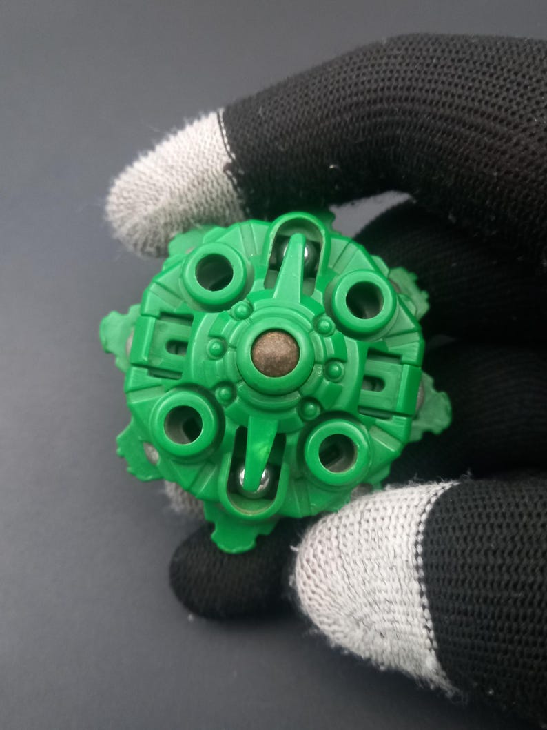 May include: A green plastic toy component with a circular design, featuring multiple holes and a central brown element. The object is held by a hand wearing a black and white glove, with a dark background.