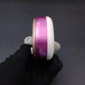 Puede incluir: Un peque&ntilde;o estuche de cosm&eacute;ticos redondo, blanco y magenta. El estuche tiene un borde dorado y una parte superior redondeada blanca. La secci&oacute;n magenta es una banda brillante en el medio. El estuche es sostenido por una mano enguantada.