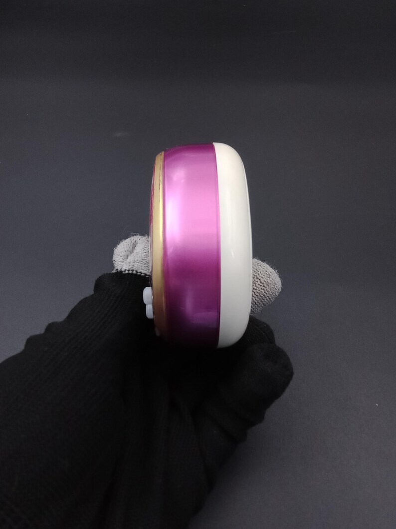 Puede incluir: Un peque&ntilde;o estuche de cosm&eacute;ticos redondo, blanco y magenta. El estuche tiene un borde dorado y una parte superior redondeada blanca. La secci&oacute;n magenta es una banda brillante en el medio. El estuche es sostenido por una mano enguantada.