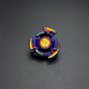 Könnte beinhalten: Ein lila, gelbes und rotes Beyblade-Spielzeug. Das Spielzeug hat ein kreisförmiges Design mit drei gelben Klingen und dem Wort "BEYBLADE" in Grün. Die Mitte zeigt eine gelb-orange Grafik.