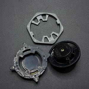 K&ouml;nnte beinhalten: Drei Metallteile f&uuml;r ein Beyblade-Spielzeug. Die Teile sind ein silberner sechseckiger Rahmen, ein schwarzer Kreisel mit silberner Mitte und ein silbernes sternf&ouml;rmiges Teil mit schwarzer Mitte.