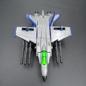 May include: A silver and blue toy jet with black missile attachments. The jet has a green cockpit and a black nose. The wings and tail have blue accents. The toy is on a dark gray surface.