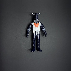 May include: A black and white action figure with a black helmet and a white chest piece. The figure has a red heart on its chest and is standing on a gray background.