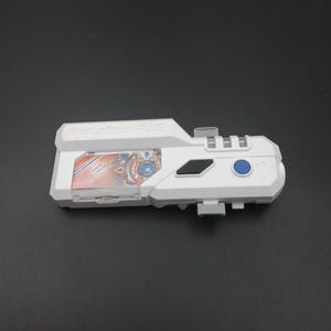 Pode incluir: Um brinquedo Beylogger PLUS branco com um botão azul e uma janela transparente exibindo um gráfico colorido. O brinquedo tem formato retangular com vários elementos salientes sobre um fundo escuro.