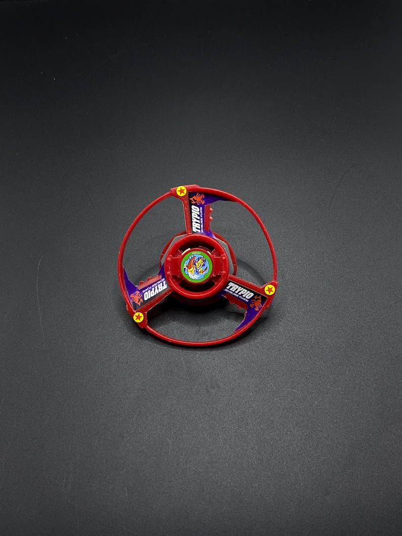 May include: Red and purple plastic toy top with a red scorpion design and the word "TRYPIO" printed on it.