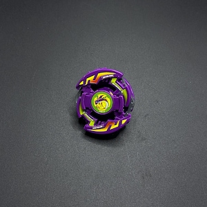 Könnte beinhalten: Lila und gelbe Beyblade mit einem Drachen-Design in der Mitte. Die Beyblade hat eine durchsichtige Plastikmitte und einen lila und gelben Plastik-Außenring.
