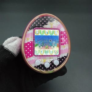 Puede incluir: Un dispositivo digital port&aacute;til rosa y negro con una pantalla que muestra una escena de juego pixelada. El dispositivo tiene tres botones blancos debajo de la pantalla y est&aacute; decorado con lunares y lazos.