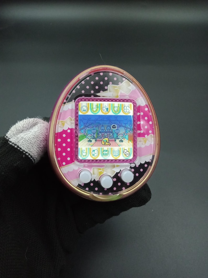 Puede incluir: Un dispositivo digital port&aacute;til rosa y negro con una pantalla que muestra una escena de juego pixelada. El dispositivo tiene tres botones blancos debajo de la pantalla y est&aacute; decorado con lunares y lazos.