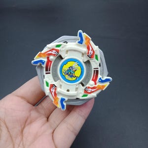 May include: A white and silver toy with colorful accents. The toy has a central blue circle with a yellow and blue illustration. The edges feature red, orange, green, and blue details. The toy is held in a hand.