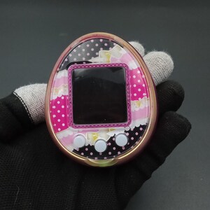 Puede incluir: Un dispositivo digital port&aacute;til rosa y dorado para mascotas con una cara estampada. La cara presenta una pantalla negra, tres botones blancos y un dise&ntilde;o con lunares rosas, flores blancas y lunares negros.