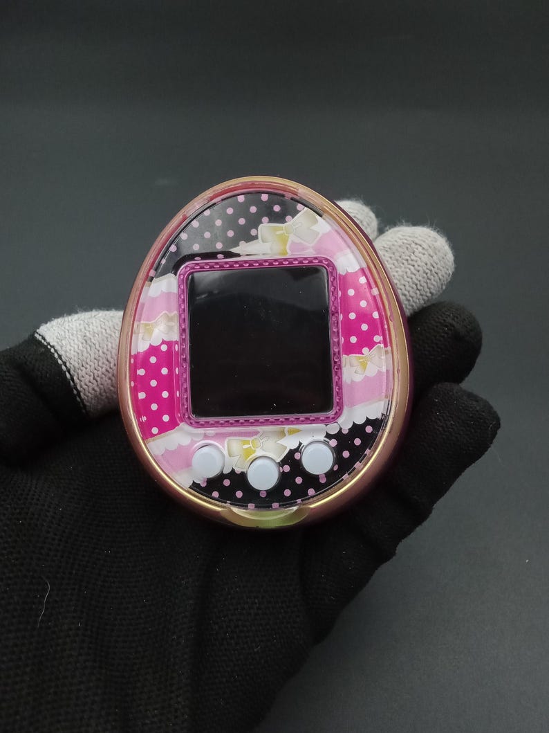 Puede incluir: Un dispositivo digital port&aacute;til rosa y dorado para mascotas con una cara estampada. La cara presenta una pantalla negra, tres botones blancos y un dise&ntilde;o con lunares rosas, flores blancas y lunares negros.