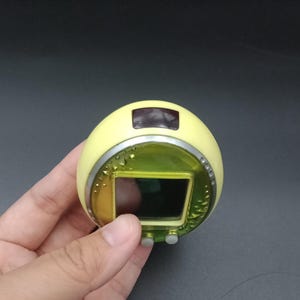 May include: A handheld, yellow electronic device with a rectangular screen and two gray buttons. The device has a silver trim and a small, dark screen at the top. The device is held in a hand against a dark background.